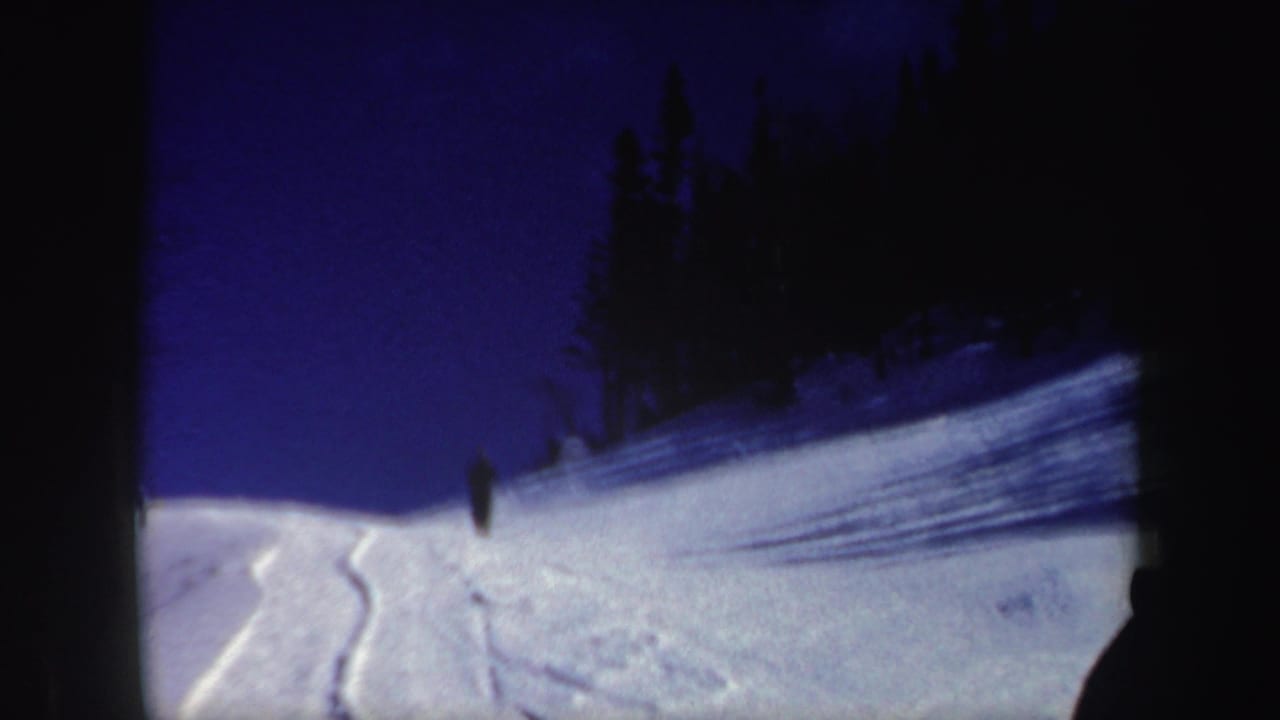 1962 New Hampshire 8mm frame showing a broad snowfield, ski tracks, dark tree silhouettes, and a small distant skier.