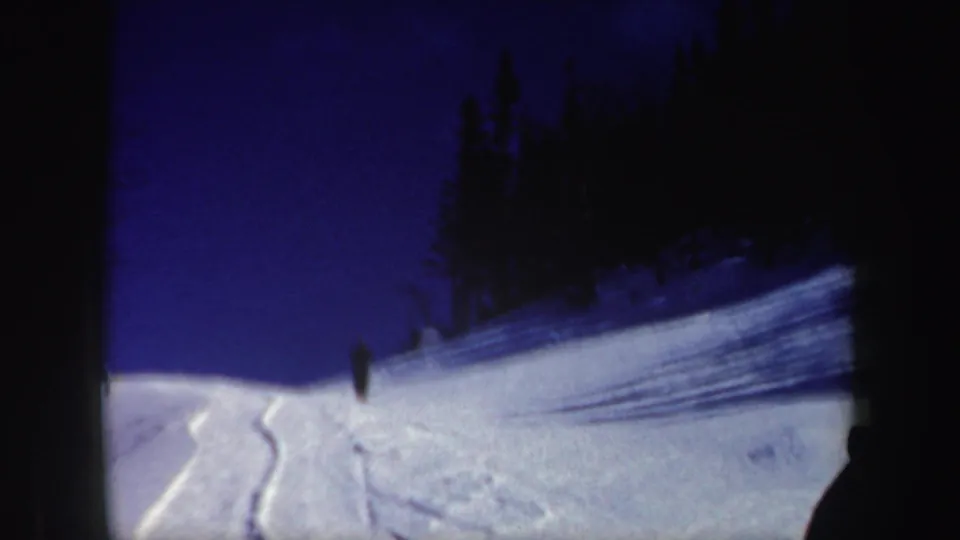 1962 New Hampshire 8mm frame showing a broad snowfield, ski tracks, dark tree silhouettes, and a small distant skier.