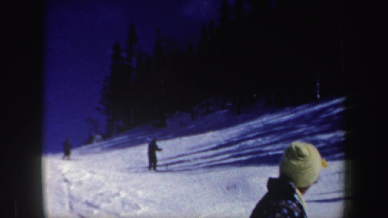 1962 New Hampshire 8mm frame showing distant skiers descending a snowy wooded slope while a person in a light knit hat watches from the foreground.