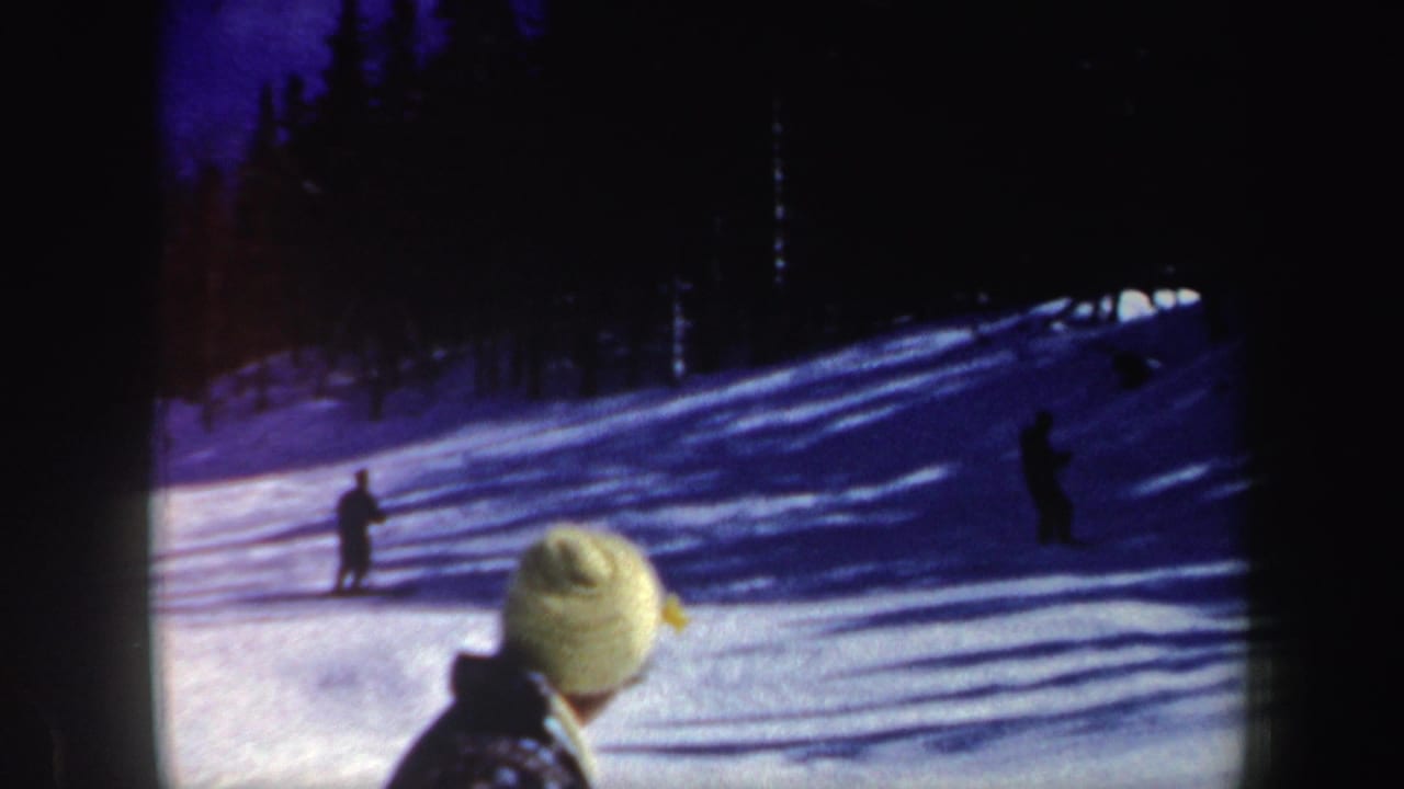 8mm home-movie frame of skiers on a shaded snowy slope in New Hampshire, with a foreground watcher partly visible at the bottom right.