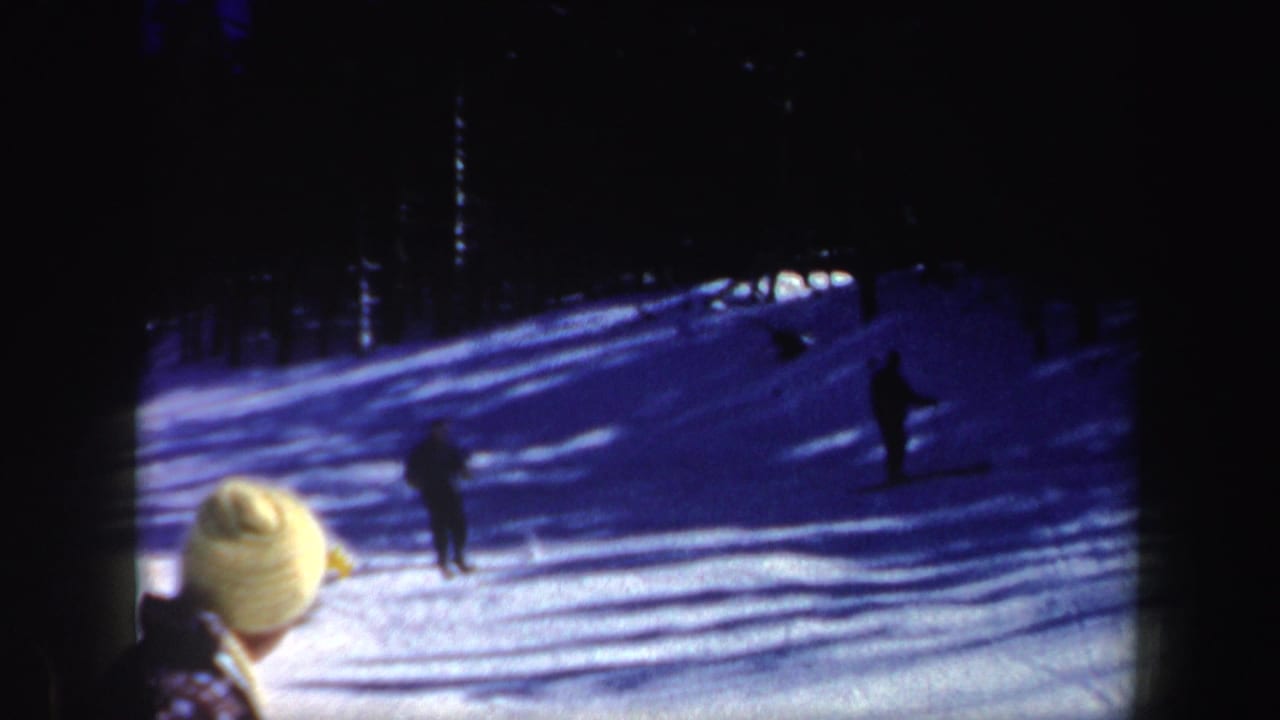 Late-frame view of two skiers on a blue-shadowed New Hampshire slope, with a partial foreground person and evergreen trees behind them.