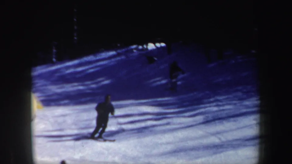 1962 New Hampshire 8mm frame showing a skier in dark clothing moving downhill across snow near a shadowed tree line.