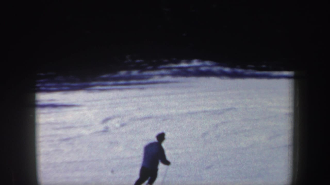 1962 New Hampshire home-movie frame with a lone skier crossing a sunlit snowfield below dark winter trees.