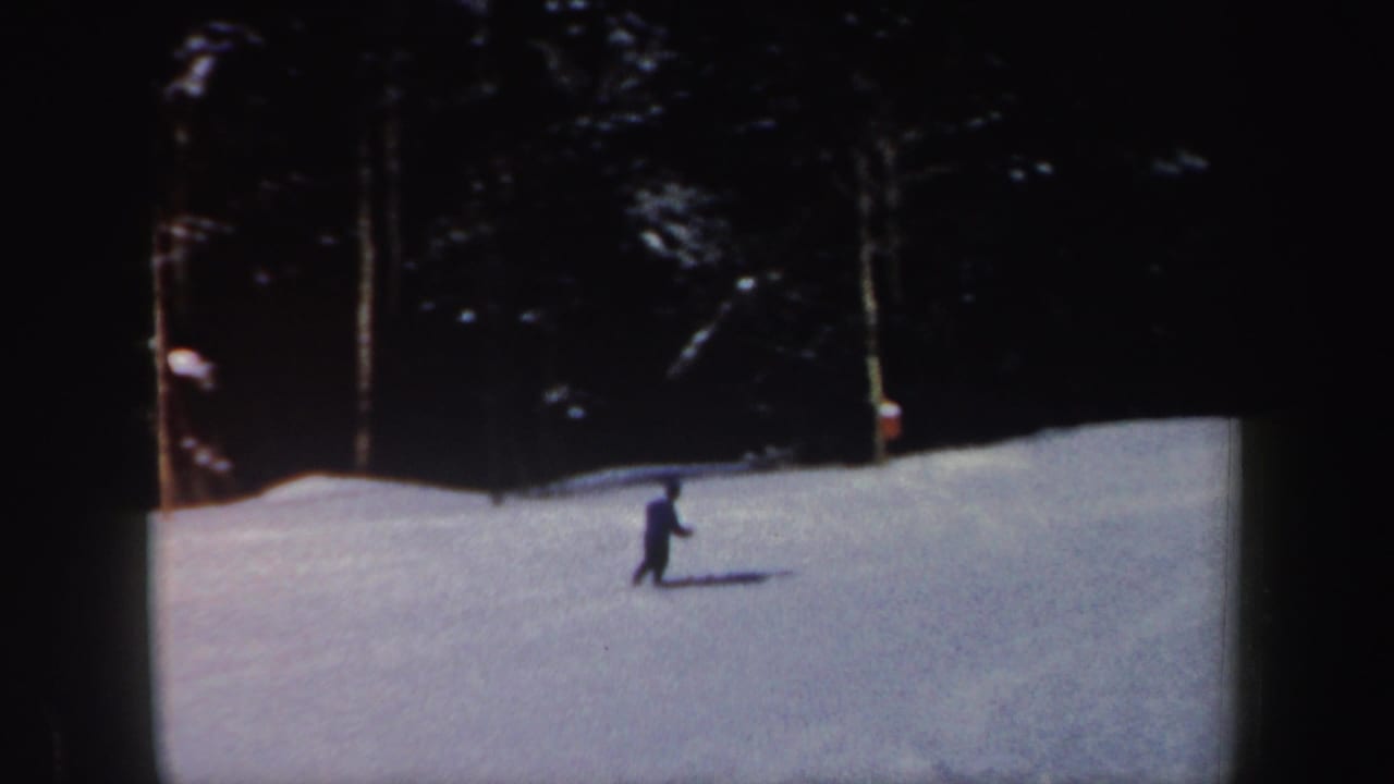 1962 New Hampshire home-movie frame with a distant skier traversing snow before a dense stand of trees.