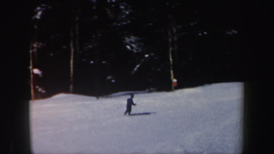 1962 New Hampshire home-movie frame with a distant skier traversing snow before a dense stand of trees.