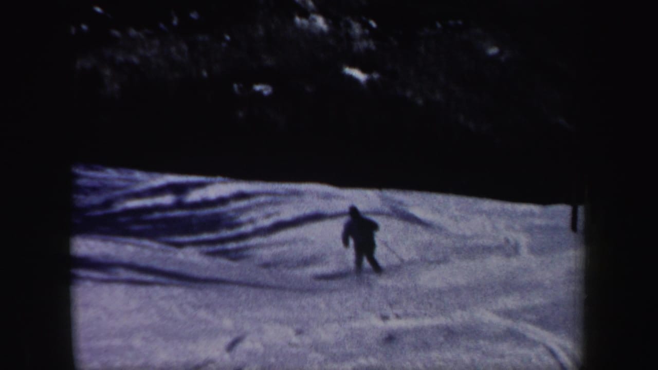 A lone adult skier continues downhill across a tracked snowy slope, framed by dark woods in 1962 home-movie footage.