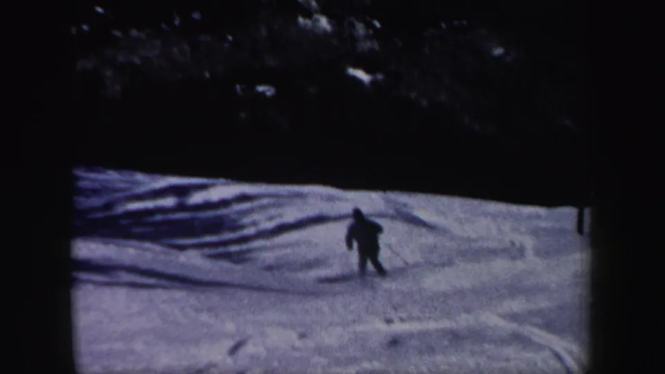 A lone adult skier continues downhill across a tracked snowy slope, framed by dark woods in 1962 home-movie footage.