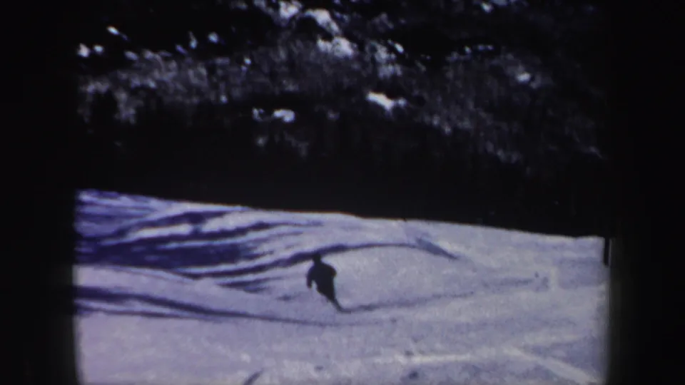 1962 New Hampshire 8mm frame showing a lone skier moving across a snowy slope with tree shadows and a dark wooded background.