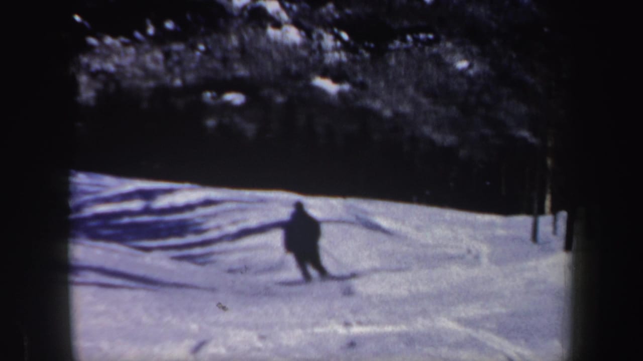 1962 New Hampshire home-movie frame of a skier in dark clothing crossing a bright snowfield below a shaded tree line.