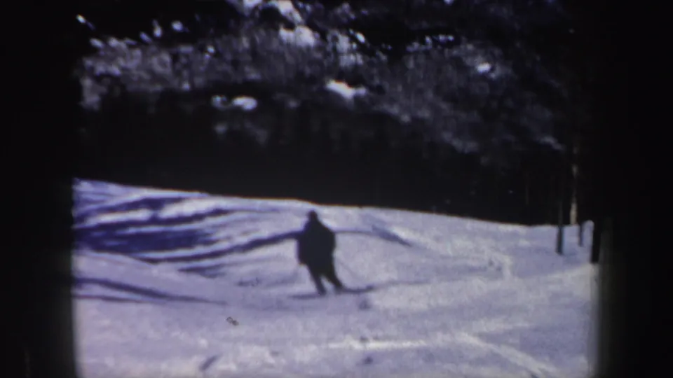 1962 New Hampshire home-movie frame of a skier in dark clothing crossing a bright snowfield below a shaded tree line.