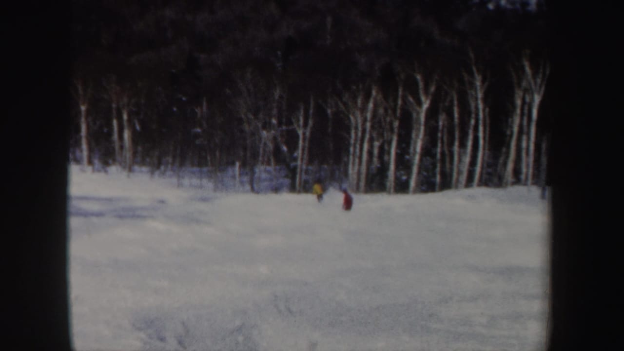 1962 New Hampshire 8mm frame with two distant skiers in red and yellow moving down a snowy wooded slope.