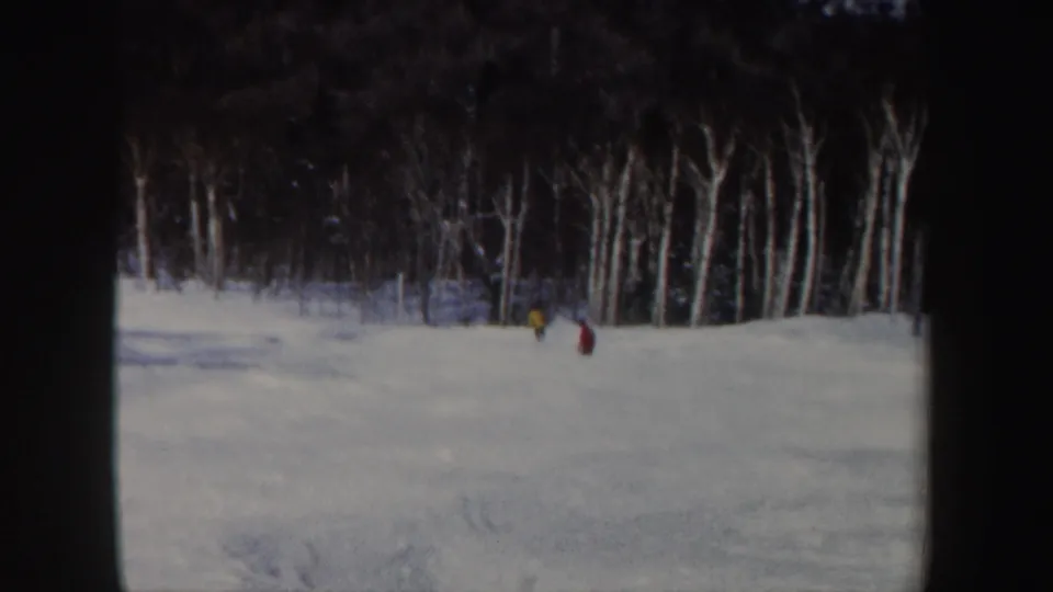 1962 New Hampshire 8mm frame with two distant skiers in red and yellow moving down a snowy wooded slope.