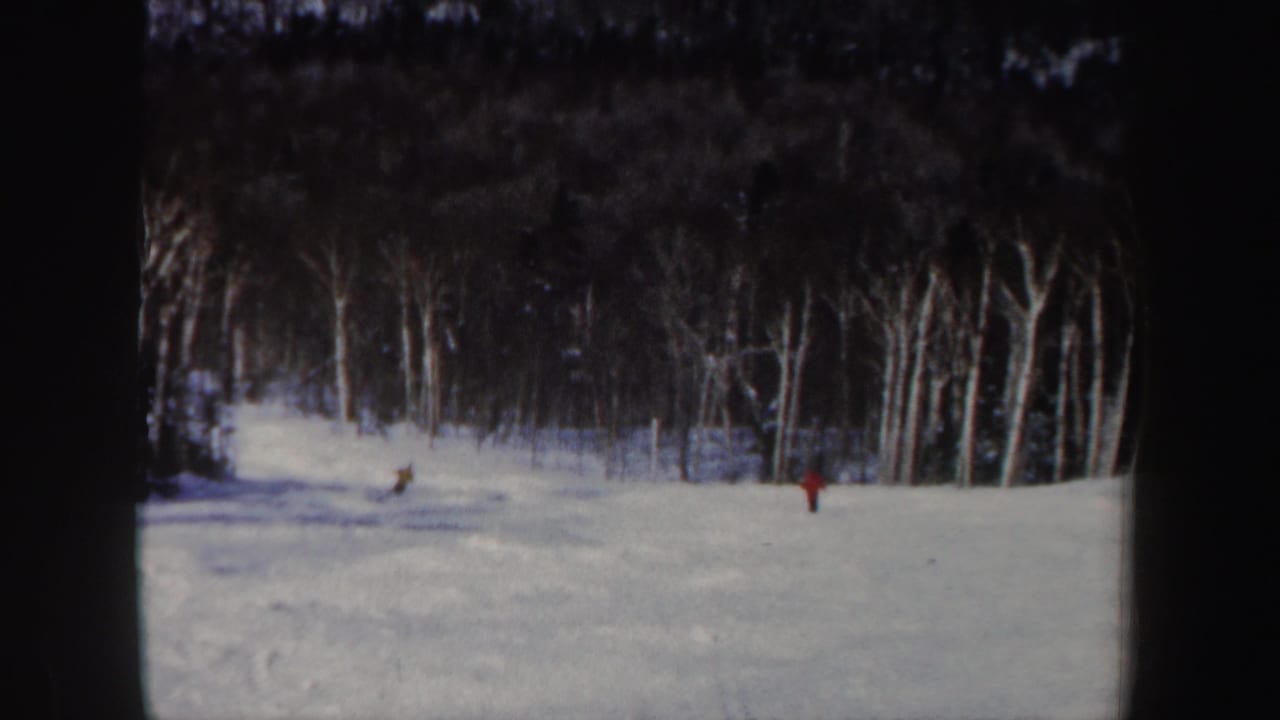 1962 home-movie frame of two distant skiers on a bright snowy slope with a dark New Hampshire forest behind them.