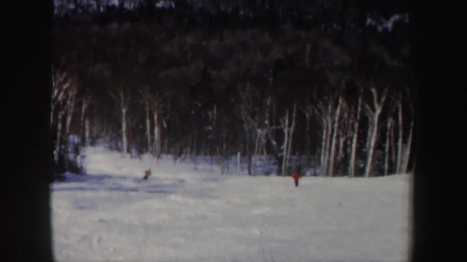 1962 home-movie frame of two distant skiers on a bright snowy slope with a dark New Hampshire forest behind them.