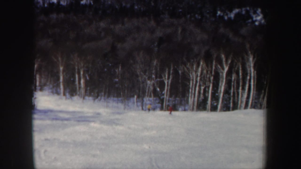 1962 New Hampshire 8mm frame showing distant skiers crossing a snowy slope bordered by bare winter trees.