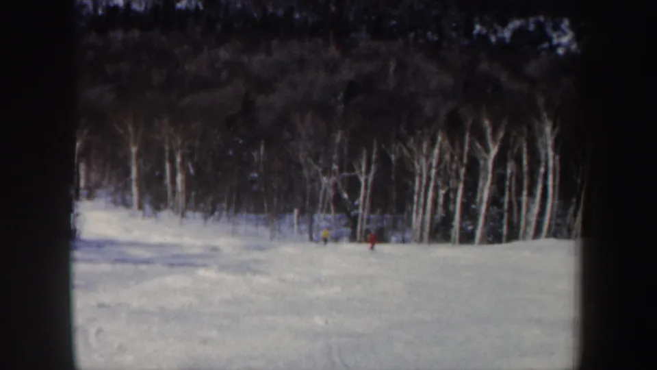 1962 New Hampshire 8mm frame showing distant skiers crossing a snowy slope bordered by bare winter trees.