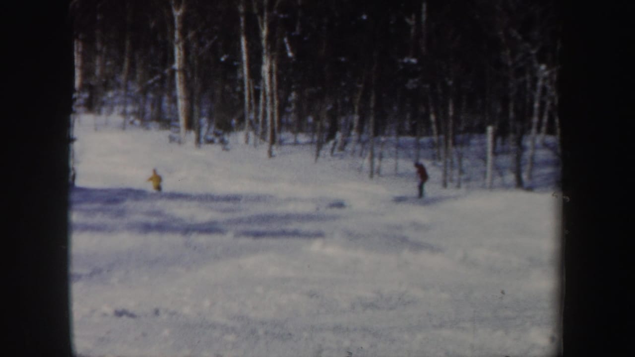 1962 New Hampshire home-movie frame with two distant skiers, including a yellow figure, on a snowy forest slope.