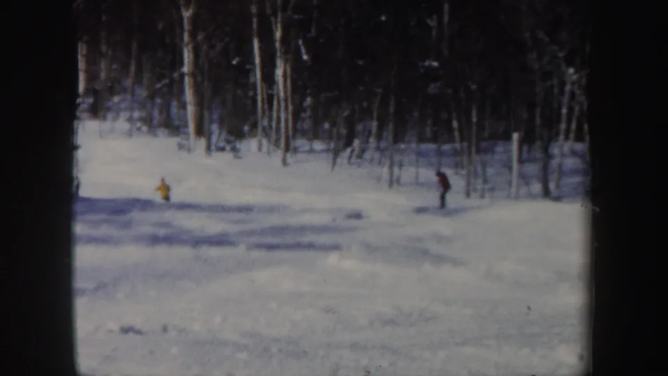 1962 New Hampshire home-movie frame with two distant skiers, including a yellow figure, on a snowy forest slope.