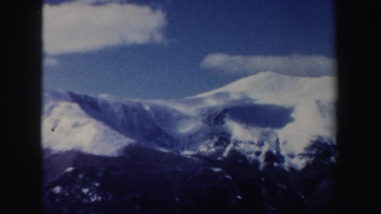 1962 New Hampshire 8mm frame of distant snow-covered peaks, dark valley shapes, blue sky, and scattered white clouds.