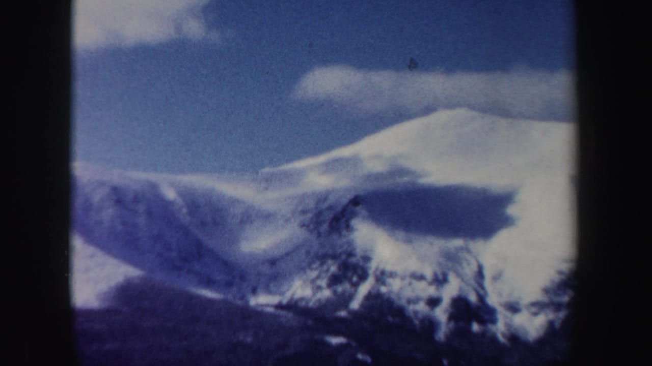 1962 New Hampshire 8mm frame showing a snow-bright ridge, dark mountain shoulder, and a low white cloud in blue sky.