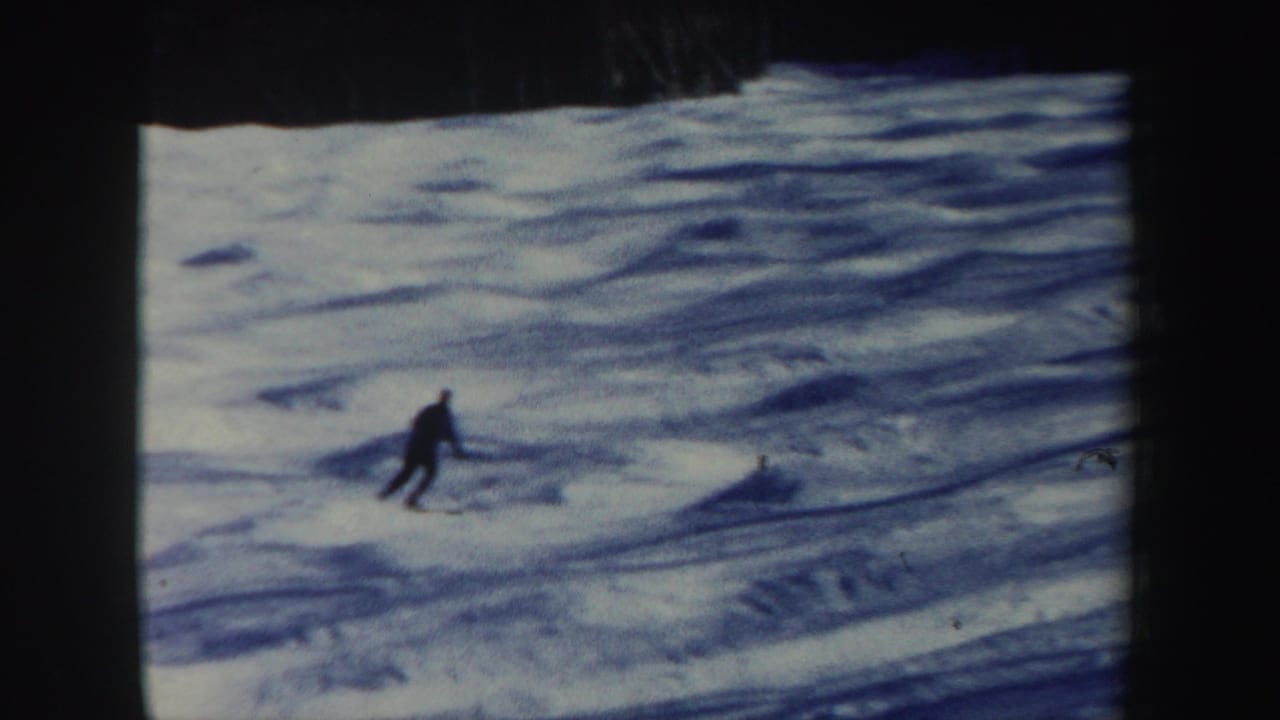 A lone skier moves across a snowy slope with dark trees at the top edge.
