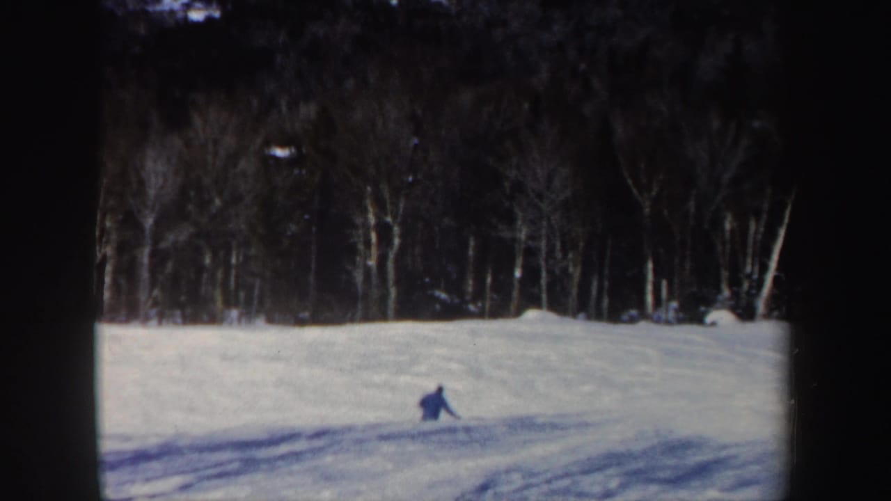 A lone skier in blue moves across a snowy New Hampshire slope in 1962, with bare trees and dark woods behind.