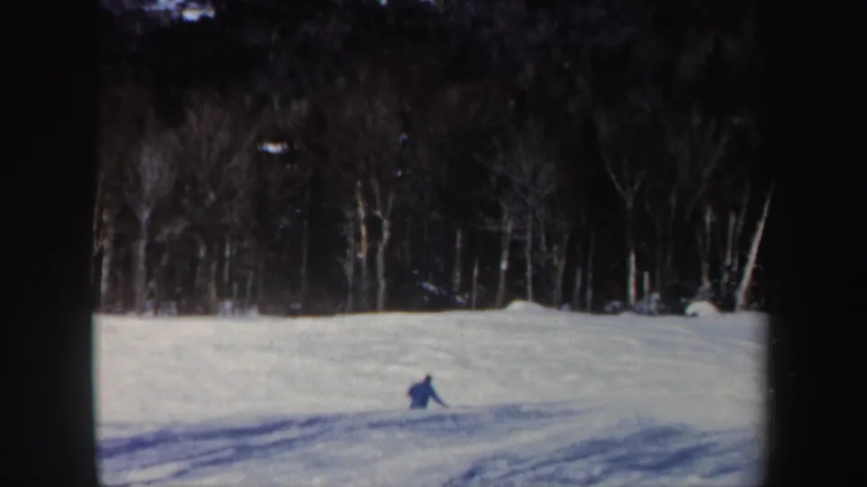 A lone skier in blue moves across a snowy New Hampshire slope in 1962, with bare trees and dark woods behind.