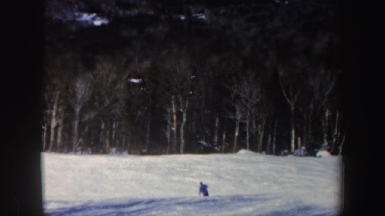 The skier appears farther across the open snowfield, framed by a dark line of leafless New Hampshire trees.