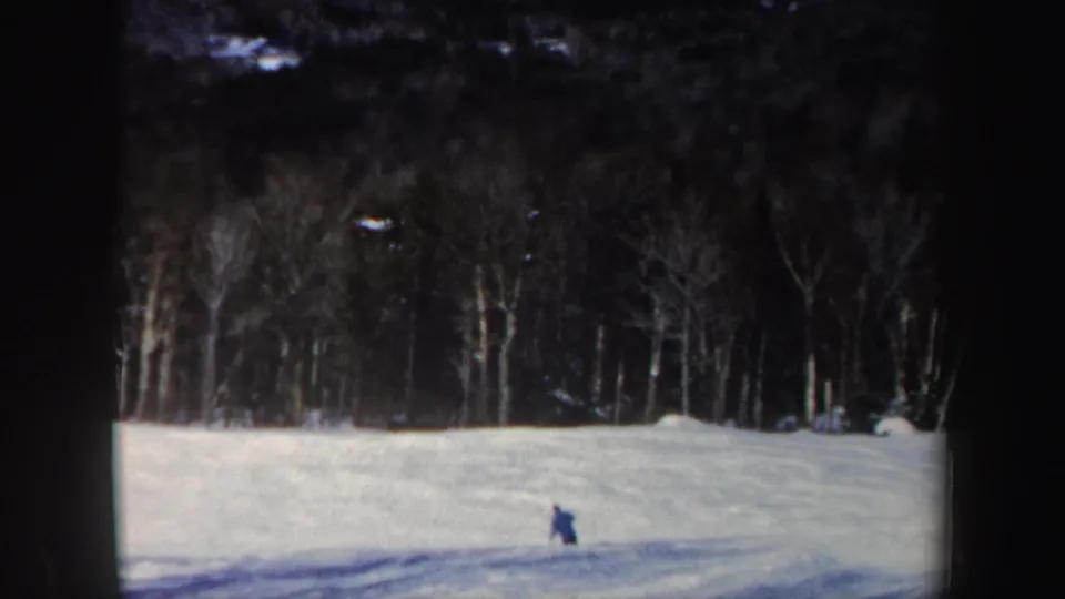 The skier appears farther across the open snowfield, framed by a dark line of leafless New Hampshire trees.