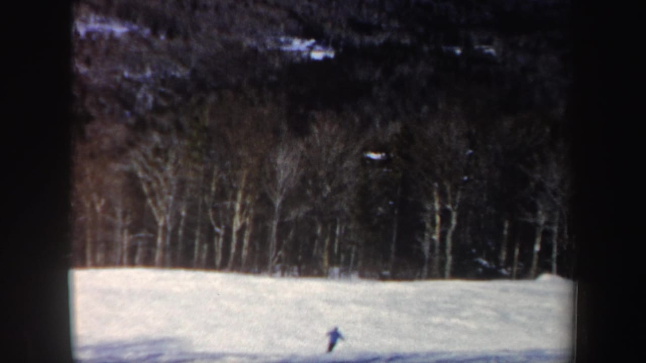 A distant skier passes near the lower slope, with uneven snow and a dense winter forest visible behind.