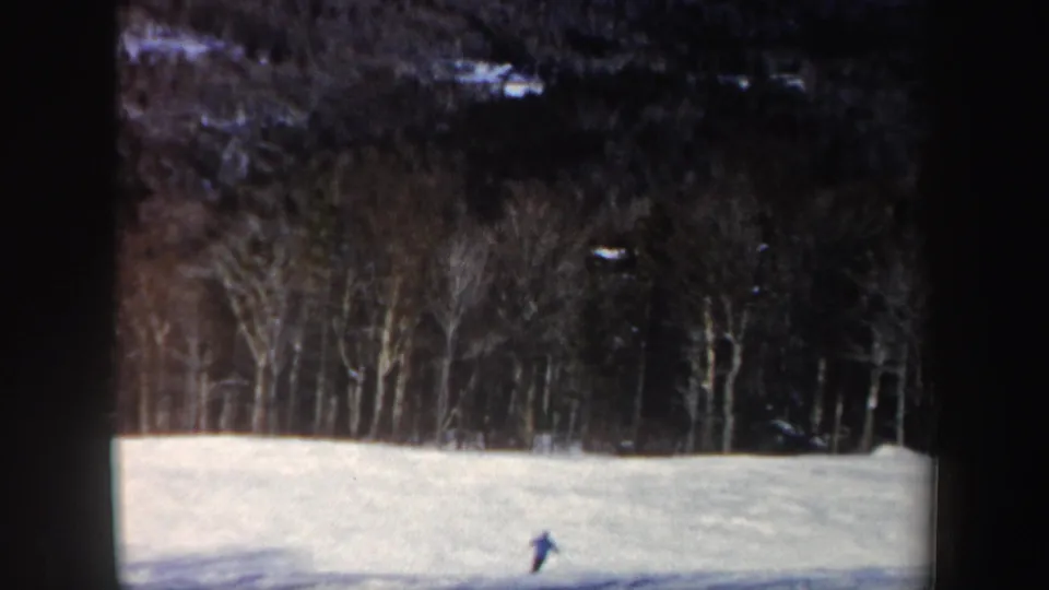 A distant skier passes near the lower slope, with uneven snow and a dense winter forest visible behind.