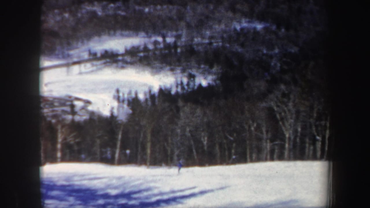 A distant skier crosses a foreground snow slope while darker New Hampshire woods and hills recede behind.