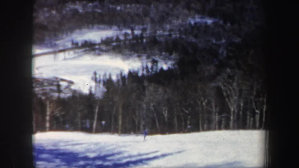 A distant skier crosses a foreground snow slope while darker New Hampshire woods and hills recede behind.