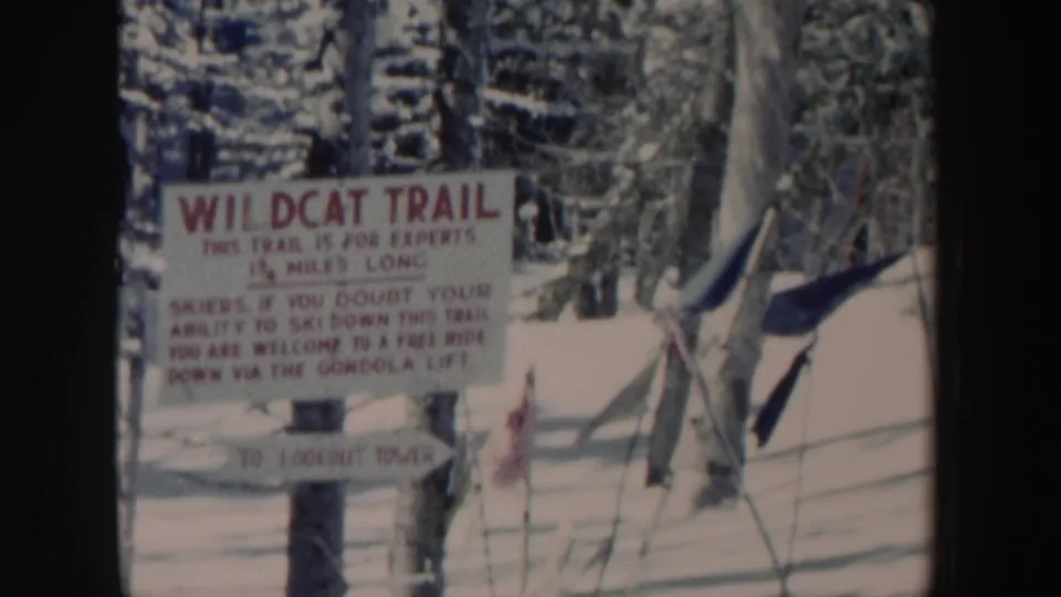 1962 New Hampshire 8mm frame showing a Wildcat Trail warning sign at a snowy ski area, with trees, fencing, and lift poles nearby.
