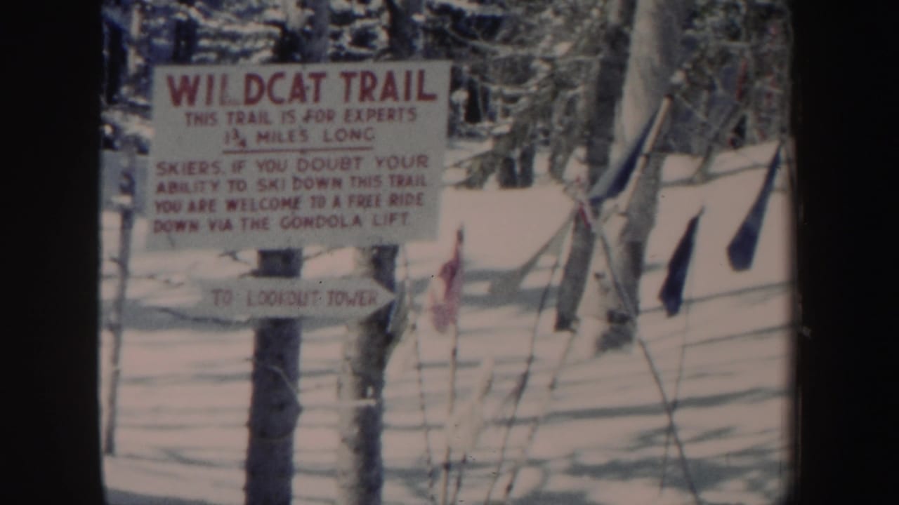 1962 New Hampshire 8mm frame showing the Wildcat Trail sign in a snowy wooded ski area, with poles, fencing, and a smaller sign below.