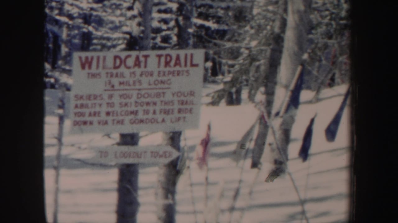 1962 New Hampshire 8mm frame of a Wildcat Trail warning sign and smaller direction sign amid snow, trees, fencing, and ski-area poles.