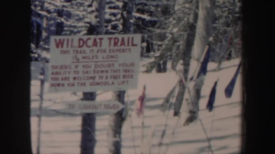 1962 New Hampshire 8mm frame of a Wildcat Trail warning sign and smaller direction sign amid snow, trees, fencing, and ski-area poles.