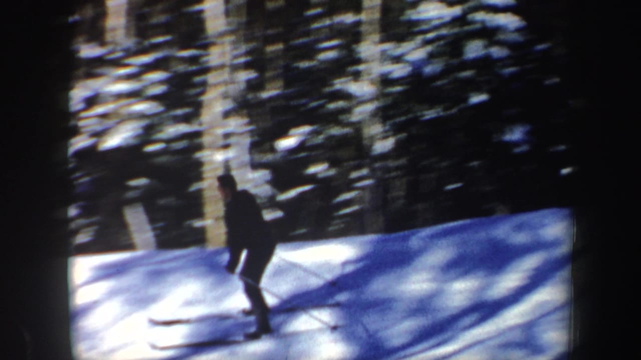 A lone skier in dark winter clothing passes through a sunlit snowy forest, filmed in New Hampshire in 1962.