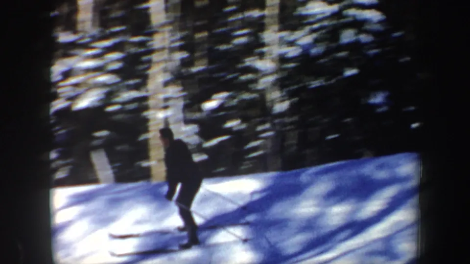 A lone skier in dark winter clothing passes through a sunlit snowy forest, filmed in New Hampshire in 1962.