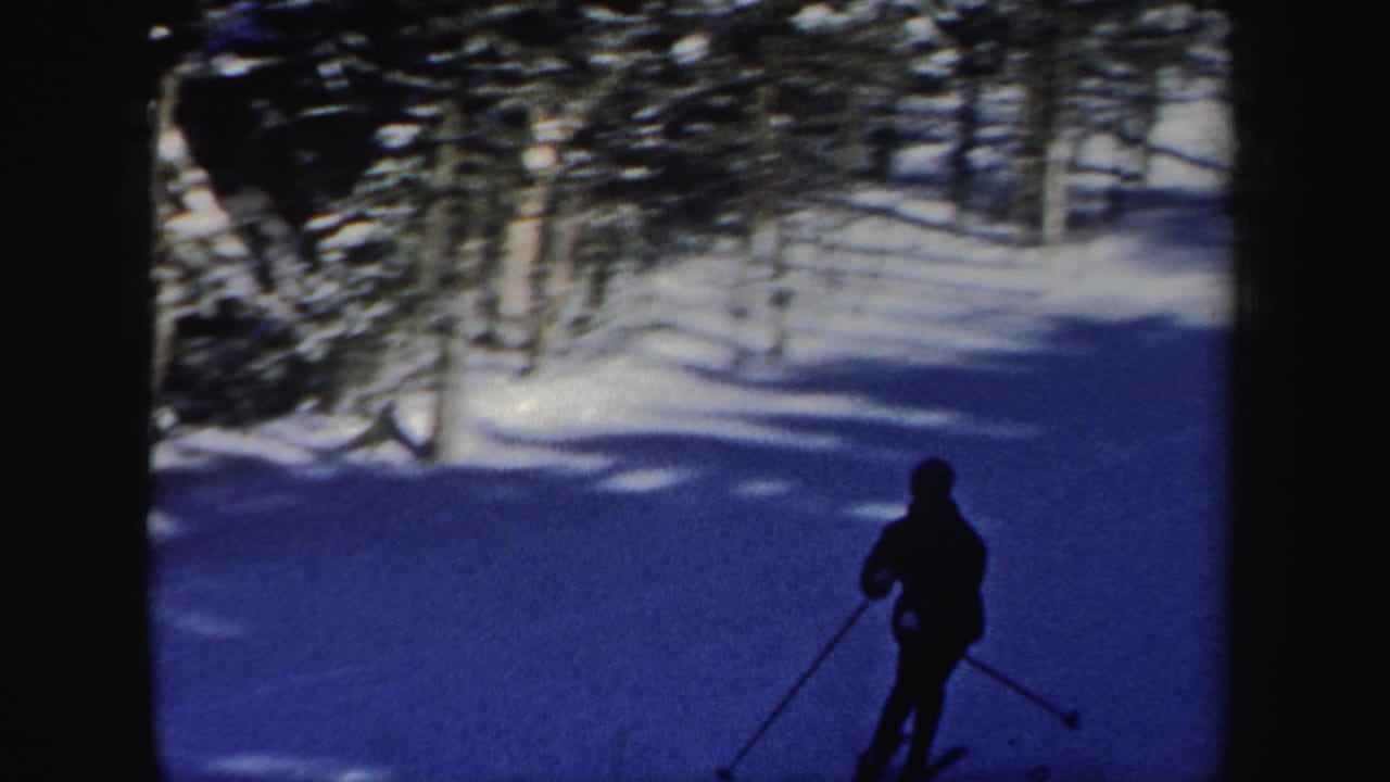 A distant skier moves down a snowy New Hampshire slope in 1962 home-movie footage, framed by dark trees and blue winter shadows.