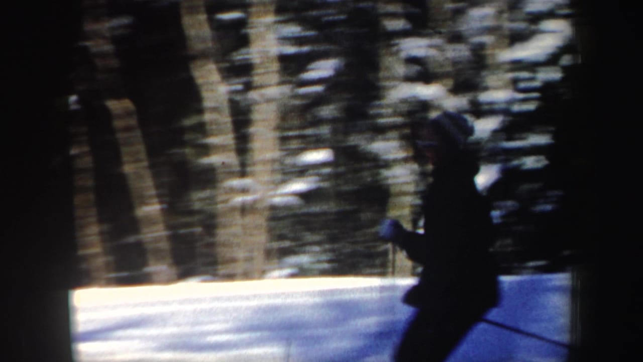 A skier in dark clothing and a striped knit hat passes across the snowy frame, with wooded background blur from handheld 8mm filming.