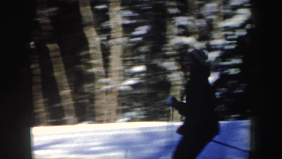 A skier in dark clothing and a striped knit hat passes across the snowy frame, with wooded background blur from handheld 8mm filming.