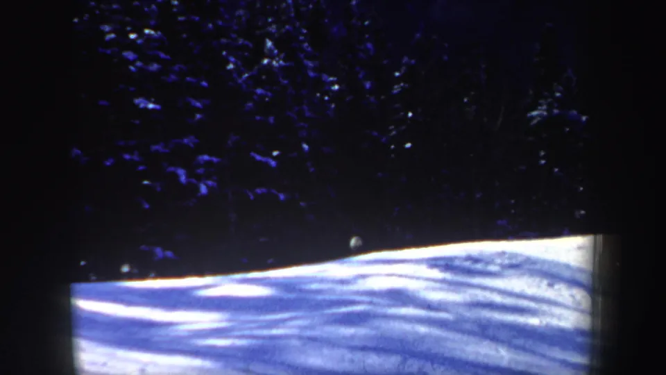 A dim 8mm frame of a snow-covered slope below shadowed evergreens, with the skier barely visible near the tree line.