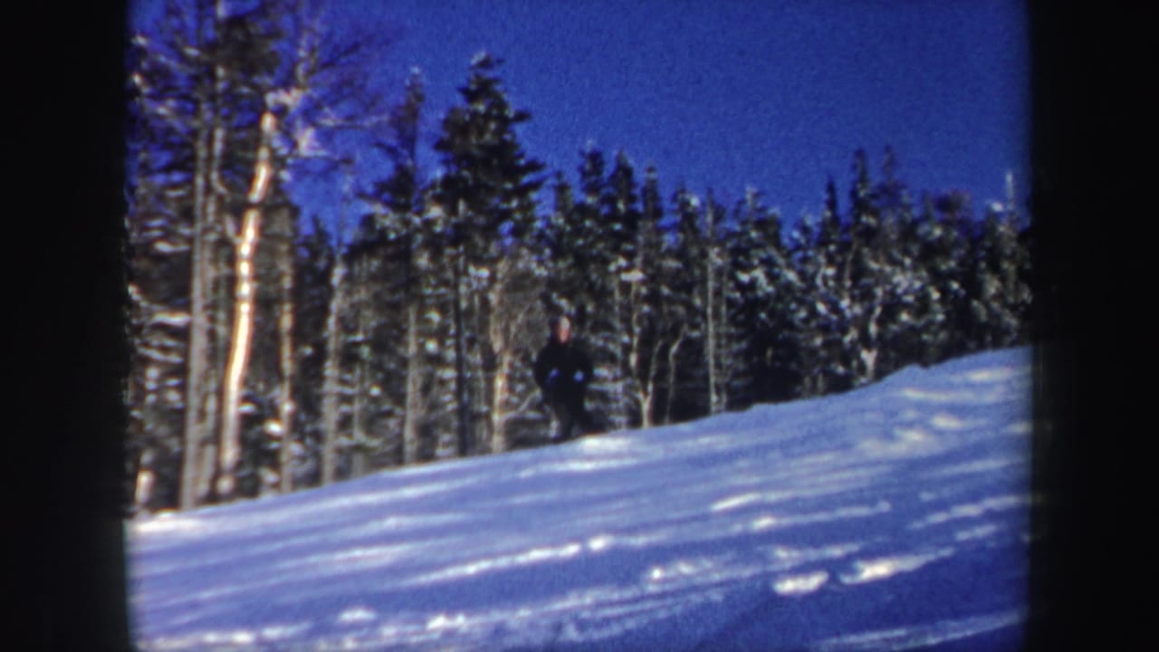 A distant skier descends a sunny New Hampshire snow slope beside mixed trees under a deep blue sky in 1962 8mm footage.