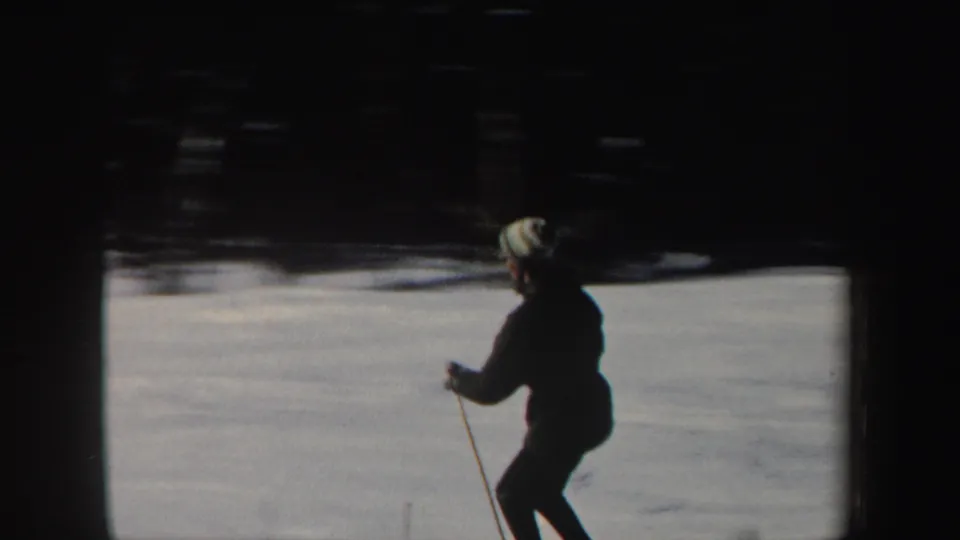 In 1962 New Hampshire home-movie footage, a skier in a dark coat and light striped hat crosses a snow-covered slope.