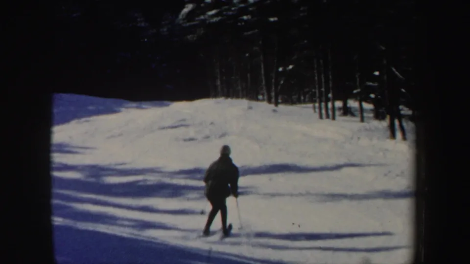 A distant skier descends a sunlit New Hampshire snow slope, with long shadows and trees along the upper edge.