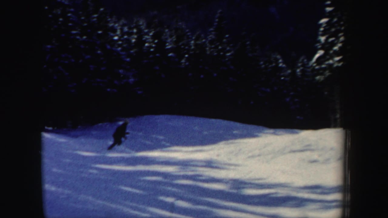 A lone skier leans into the descent on a snowy wooded slope, captured in soft 8mm color home-movie footage.