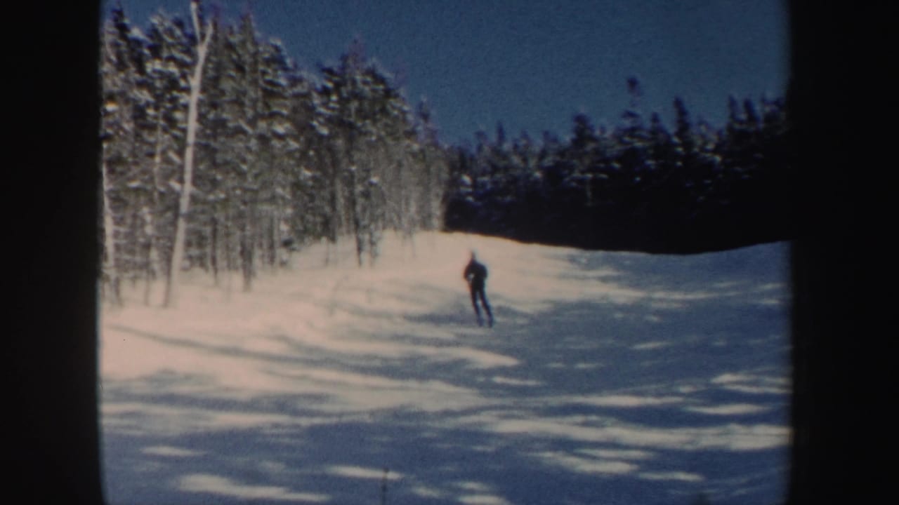 A lone skier in dark clothing crosses a sunlit snowy slope in New Hampshire archival home-movie footage.