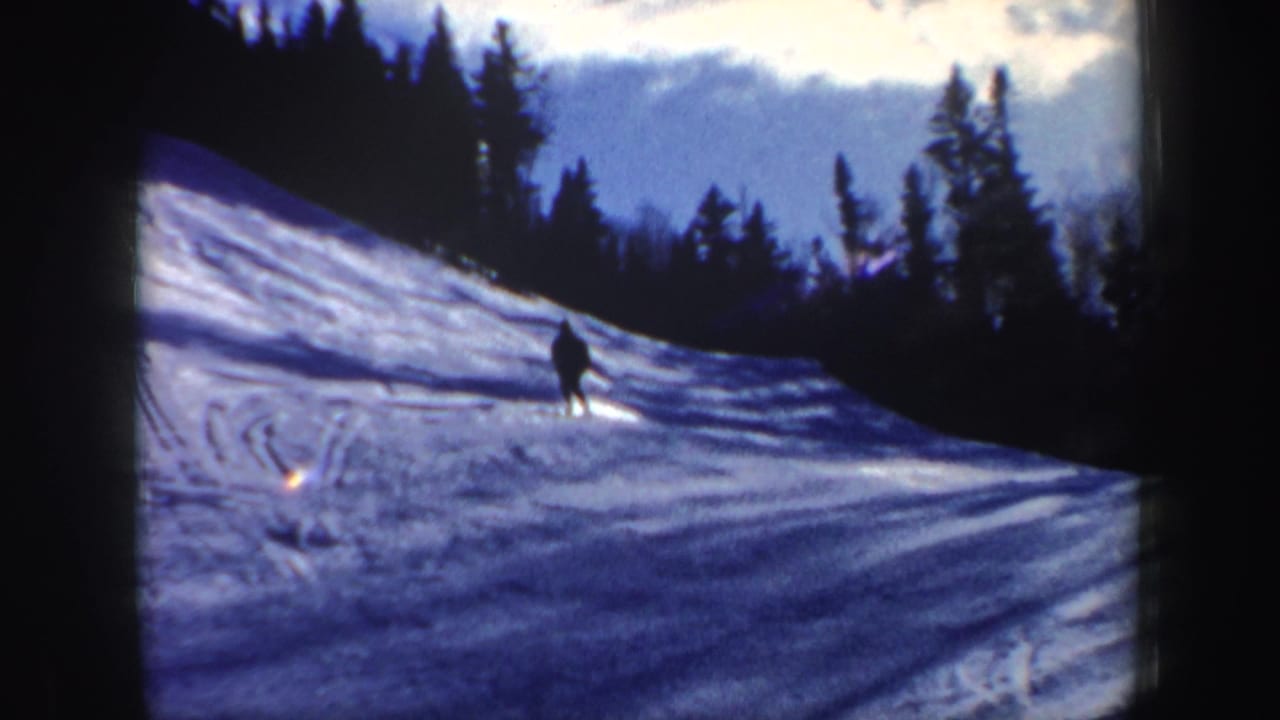 1962 New Hampshire frame showing one skier descending a tracked snow slope beneath a forested ridge and blue sky.