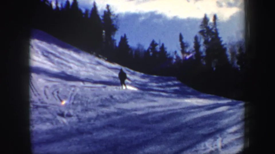 1962 New Hampshire frame showing one skier descending a tracked snow slope beneath a forested ridge and blue sky.